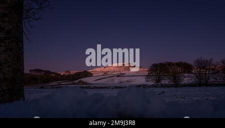 Nachtfoto von Pen-y-ghent im Yorkshire Dales National Park, wo der letzte Sonnenstrahl auf dem schneebedeckten Berggipfel mit Sternen zu sehen ist Stockfoto