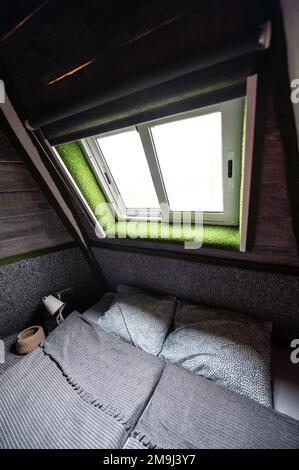 Doppelbett auf Teppichboden im Schlafzimmer der Holzkabine. Pyramidenförmiger Bungalow von innen. Kleines Zimmer, abgewinkelte Wände, Fensterbeleuchtung, Bett gemacht. Handgefertigt Stockfoto