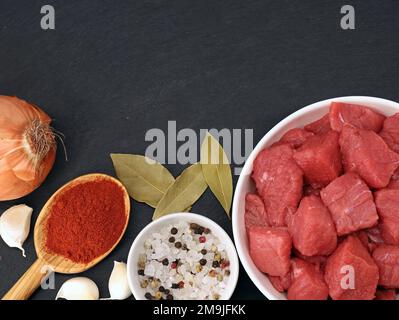 Rohes gewürfeltes Rindfleisch mit Zwiebeln, Knoblauch und Gewürzen auf einem schwarzen Steinbrett, Draufsicht der Zutaten für Gulasch Stockfoto