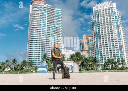 Hipster Mann mit Gepäcktasche auf Rädern stehen am Südstrand in miami. Sonniger Tag, Reisekonzept Stockfoto