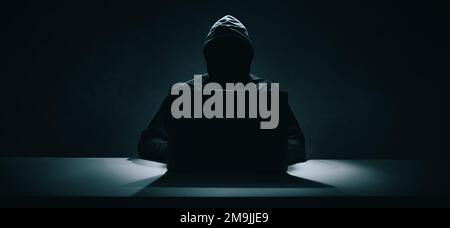 Hacker in schwarzem Hoodie, der im Dunkeln an einem Laptop arbeitet. Cybersicherheit. Banner Stockfoto