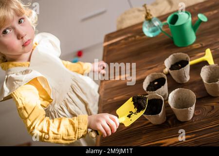 Das Kind gießt die Erde in den Topf, um Samen zu Pflanzen. Anbau von Setzlingen zur Verpflanzung im Frühjahr auf einem Ackerfeld. Stockfoto