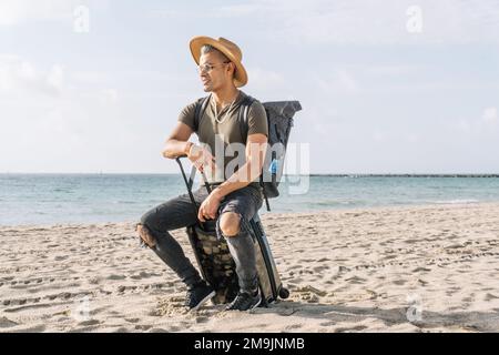 Nahaufnahme eines verlorenen Mannes, der auf seinem kleinen Koffer sitzt und das Meer am Strand sieht, bereit für den Urlaub. Stockfoto