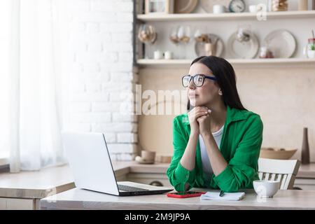 Verärgerte junge Frau, die zu Hause in der Küche vor einem Laptop sitzt. Geben Sie auf der Tastatur ein, suchen Sie, überprüfen Sie. Sie schaut durchdacht aus dem Fenster und legt ihren Kopf auf ihre Hände. Stockfoto