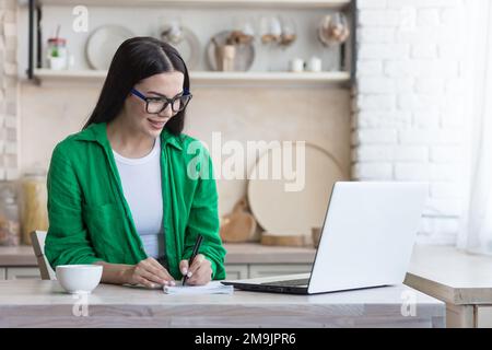 Eine junge Studentin, eine Freiberuflerin, arbeitet online, ferngesteuert von einem Laptop zu Hause. Er sitzt am Tisch, hört dem Vortrag aufmerksam zu, schreibt mit einem Stift in einem Notizbuch. Stockfoto