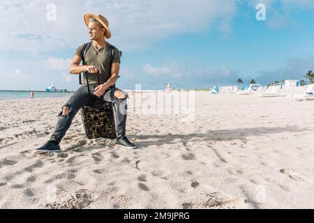 Gut aussehend und fit Kerl posiert mit einem kleinen Koffer. Ein Mann am Strand, der das Meer betrachtet. Reisekonzept Stockfoto