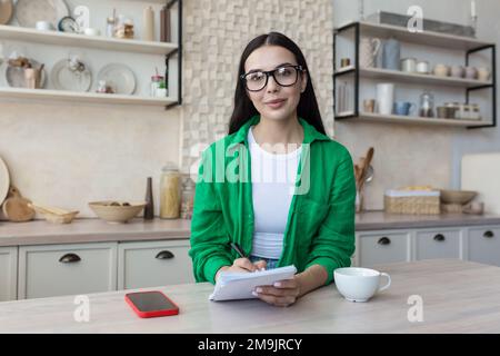 Porträt einer jungen Brillenlehrerin, die zu Hause in der Küche vor der Kamera sitzt. Arbeitet von zu Hause aus online. Er hält ein Notizbuch mit einem Stift in der Hand und lächelt. Stockfoto
