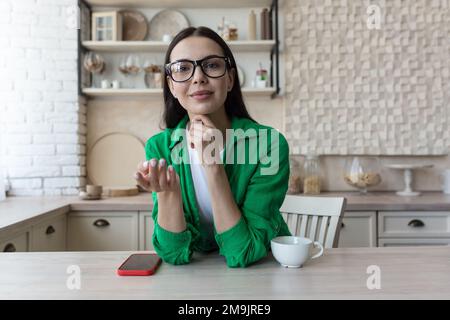 Eine junge, wunderschöne Bloggerin sitzt zu Hause in der Küche und spricht online mit der Kamera, erklärt mit ihren Händen und lächelt. Stockfoto