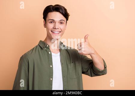 Foto eines zufriedenen, gut gelaunten Mannes mit braunem Haar gekleidetem Khaki-Hemd, das den Daumen nach oben zeigt, wie genehmigt isoliert auf beigefarbenem Hintergrund Stockfoto