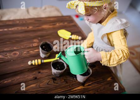 Wintervorbereitung für die Aussaat. Ein Mädchen gießt die Samen in Töpfen auf einem Holztisch mit gesäten Samen. Setzlinge für den Garten anbauen. Vegeta Stockfoto