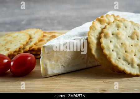 Nahaufnahme einer Scheibe französischen Brie Weichkäses mit weißer Schimmelrinde serviert mit reifen roten Pflaumentomaten und trockenen dünnen Crackern Stockfoto