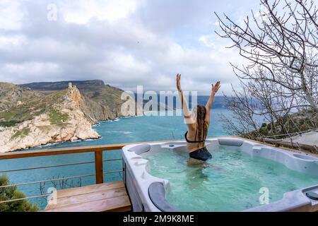 Nehmen Sie sich Zeit für sich. Badesachen im Freien mit Blick auf die Berge und das Meer. Eine Frau im schwarzen Badeanzug entspannt sich im Hotelpool und bewundert die Aussicht Stockfoto