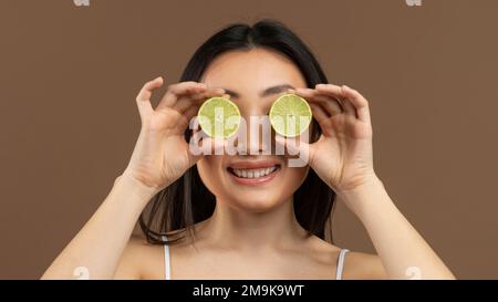 Vitamine für Schönheit und Hautpflege. Asiatische Dame, die die Augen mit Limettenhälften verdeckt, über braunem Hintergrund steht, Panorama Stockfoto