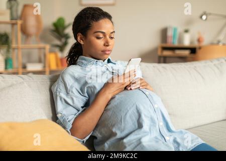 afroamerikanische Schwangere, die ihr Smartphone benutzt, zu Hause auf dem Sofa sitzt, im Internet surft oder mit einem Arzt schreibt Stockfoto