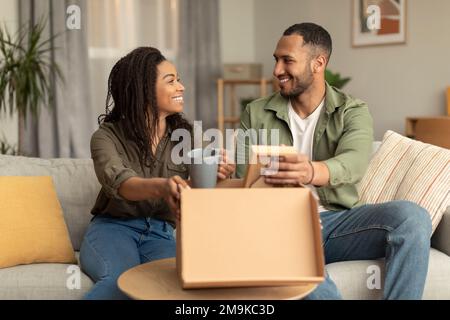 Glücklicher schwarzer Familienvater, Ehemann und Ehefrau, die Sachen aus dem Karton auspacken, nachdem sie umgezogen sind, sitzen auf dem Sofa Stockfoto