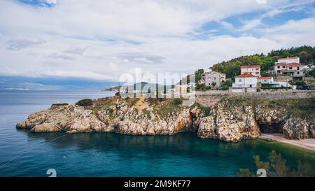 Ein Bild einer felsigen Küste der Insel Krk mit mehreren Gebäuden umgeben von Bäumen in Vrbnik, Kroatien Stockfoto