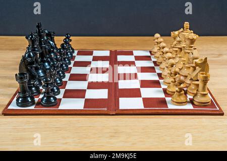 Kleines Schachset und Schachbrett, wie unten in der Mitte des Spielbretts dargestellt, mit dunklen Teilen auf der einen und leichten Teilen auf der anderen Seite. Stockfoto