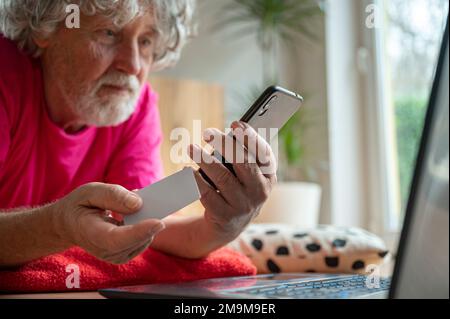 Ein älterer Mann, der auf dem Boden eines Wohnzimmers lag und ein Mobiltelefon benutzte, um einen Online-Kauf mit einer Kreditkarte zu tätigen. Stockfoto