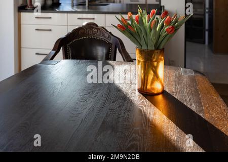 Auf einem riesigen dunklen Holztisch in den Sonnenstrahlen des Morgens steht eine gelbe Vase mit roten Tulpen. Stockfoto