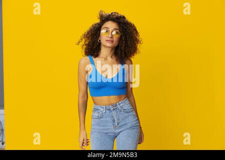 Eine Frau mit lockigem Afro-Haar in einem blauen T-Shirt auf gelbem Hintergrund tanzt fliegende Haare mit gelber Sonnenbrille, Handzeichen, schauen Sie in die Kamera Stockfoto