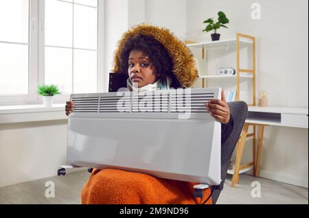 Junge gefrorene Frau, die in einem Wintermantel auf einem Stuhl zu Hause sitzt und einen elektrischen Heizkörper hält. Stockfoto
