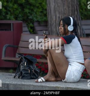 Ein Mädchen mit Kopfhörern sitzt auf der Straße und schaut mit einer Tasche in ihr Smartphone Stockfoto