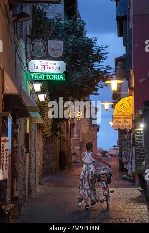 Eine Frau läuft mit dem Fahrrad eine leere, enge Straße entlang, die von Restaurantschildern in Sermione, Italien, beleuchtet wird Stockfoto