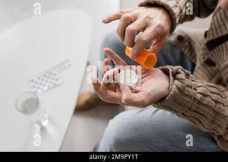 Partielle Ansicht von Mann mit parkinson-Krankheit und Tremor in Händen halten Pillen Behälter in der Nähe verschwommenes Glas Wasser, Stock Bild Stockfoto