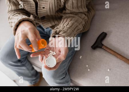 Freigestellte Ansicht eines alten Mannes, der an Parkinsonismus leidet und Behälter mit Pillen hält, während er auf dem Sofa sitzt, Stockbild Stockfoto