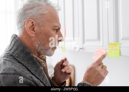 Seitenansicht eines nachdenklichen Mannes mit alzheimer-Syndrom, der auf verschwommene Haftnotiz in der Küche zeigt, Stockbild Stockfoto