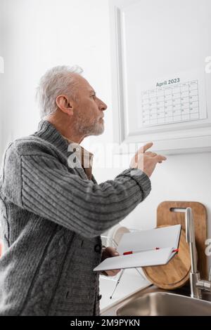 Senior Mann mit alzheimer Krankheit hält leere Notizbuch und zeigt auf Kalender in der Küche, Stock Image Stockfoto