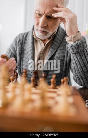 Rücksichtsvoller Senior-Mann mit Gedächtnisverlust und Blick auf Schachfigur auf verschwommenem Vordergrund, Stock-Bild Stockfoto