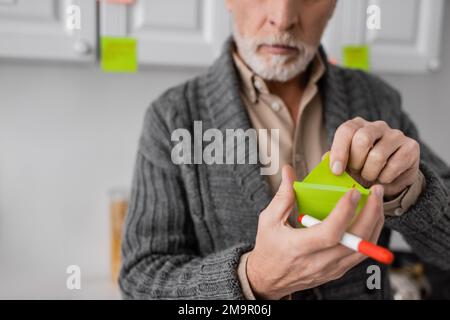 Teilansicht eines verschwommenen Seniorenmannes, der an alzheimer-Syndrom leidet und Filzstift und Haftnotizen in der Küche hält, Stockbild Stockfoto