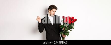 Bild von eleganten und frechen Mann in schwarzem Anzug, selbstbewusst und hält Strauß von roten Rosen, gehen auf ein romantisches Date, stehen gegen weiß Stockfoto