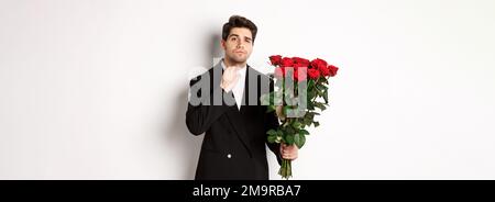 Bild von eleganten und frechen Mann in schwarzem Anzug, selbstbewusst und hält Strauß von roten Rosen, gehen auf ein romantisches Date, stehen gegen weiß Stockfoto