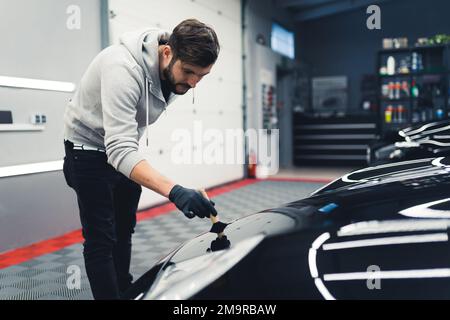 Männlicher Mechaniker, der einen Pinsel für Autodetails an seinem schwarzen Sportwagen benutzt. Professionelle Wartung eines Autos. Werkstattinnenraum. Hochwertiges Foto Stockfoto