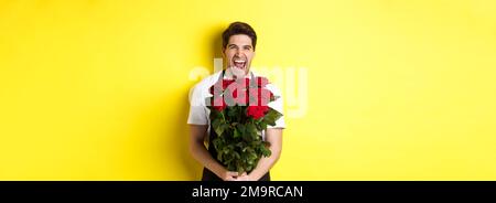 Lustiger junger Verkäufer in schwarzer Schürze, der Strauß Rosen hält, Blumenhändler lachend und über gelbem Hintergrund stehend Stockfoto