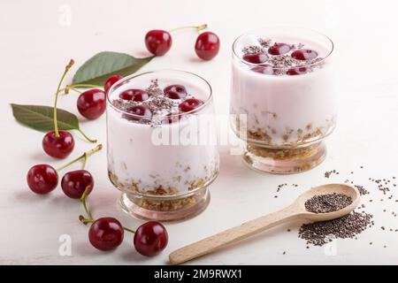 Joghurt mit Kirschen, Chiasamen und Müsli im Glas mit Holzlöffel auf weißem Holzhintergrund. Seitenansicht, Nahaufnahme. Stockfoto