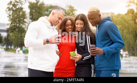 Multi-ethnische Gruppe Teenager-Freunde suchen Smartphone-Bildschirm. afroamerikanisch asiatisch kaukasisch Student verbringen Zeit zusammen multirassische Freundschaft lächelnde Menschen bunte Sportbekleidung Kaffee trinken Stockfoto