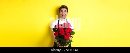 Verkäufer in Blumengeschäft trägt schwarze Schürze, geben Strauß von Rosen und lächelnd, stehend über gelbem Hintergrund Stockfoto