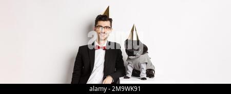 Bild von schönen jungen Mann Geburtstag feiern mit niedlichen schwarzen Mops in Party-Kostüm und Kegel auf dem Kopf, stehen über weißem Hintergrund Stockfoto