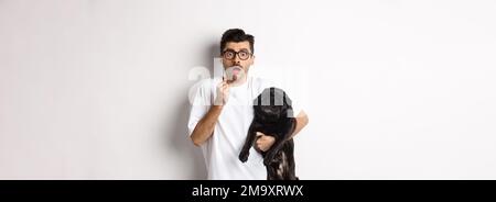 Erschrockener Hipster umarmt seine niedlichen schwarzen Welpen und starrte auf Kamera erschrocken. Hundebesitzer schaut schockiert, hält Mops, steht auf weißem Hintergrund Stockfoto