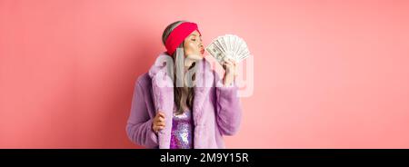 Shopping- und Modekonzept. Modische und reiche ältere Frau küssen Dollar Geld, zufrieden, tragen lila Kunstpelz Mantel mit Party-Kleid Stockfoto