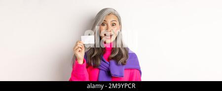 Einkaufskonzept. Glücklich asiatische alte aldy suchen beeindruckt und zeigt Kunststoff-Kreditkarte ihrer Bank, über weißem Hintergrund stehen Stockfoto