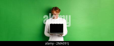 Glücklicher junger Mann mit roten Haaren, zeigt leeren Laptop-Bildschirm und zufrieden, lächelnd, während starrte auf Computer, grüner Hintergrund Stockfoto