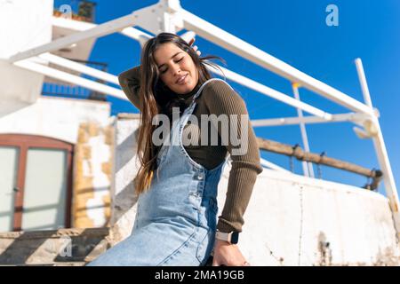 Eine ethnische Frau mit niedrigem Winkelgehalt in Denim, die Haare mit geschlossenen Augen berührt, während sie in der Nähe des alten Piers im Sonnenlicht sitzt Stockfoto