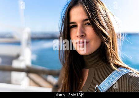 Eine attraktive, langhaarige hispanische Dame, die in der Sonne auf den unscharfen Hintergrund eines Seepiers in Seenähe in die Kamera schaut Stockfoto