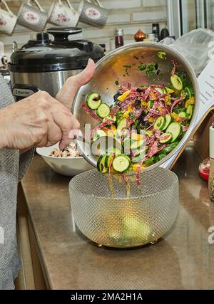 Der Koch gibt den Salat mit Zucchini, Orangen und Salami aus der Schüssel in die Salatschüsseln. Französische Gourmetküche, Holzhintergrund. Stockfoto