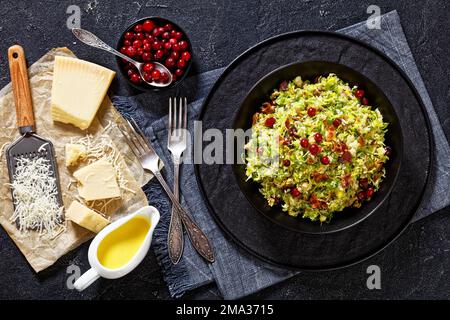 rosenkohl mit Mandeln, knusprig gebratenem Speck, geriebenem Parmesan und Preiselbeeren in schwarzer Schale auf Betontisch, horizontale Ansicht von Stockfoto