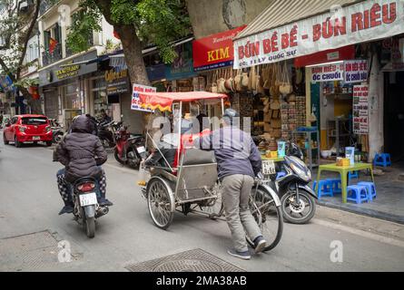 Ein traditioneller Cyclo-Pedicab-Fahrer schiebt seinen Cyclo mit zwei Passagieren auf einen leichten Hügel in Hanoi, Vietnam. Stockfoto